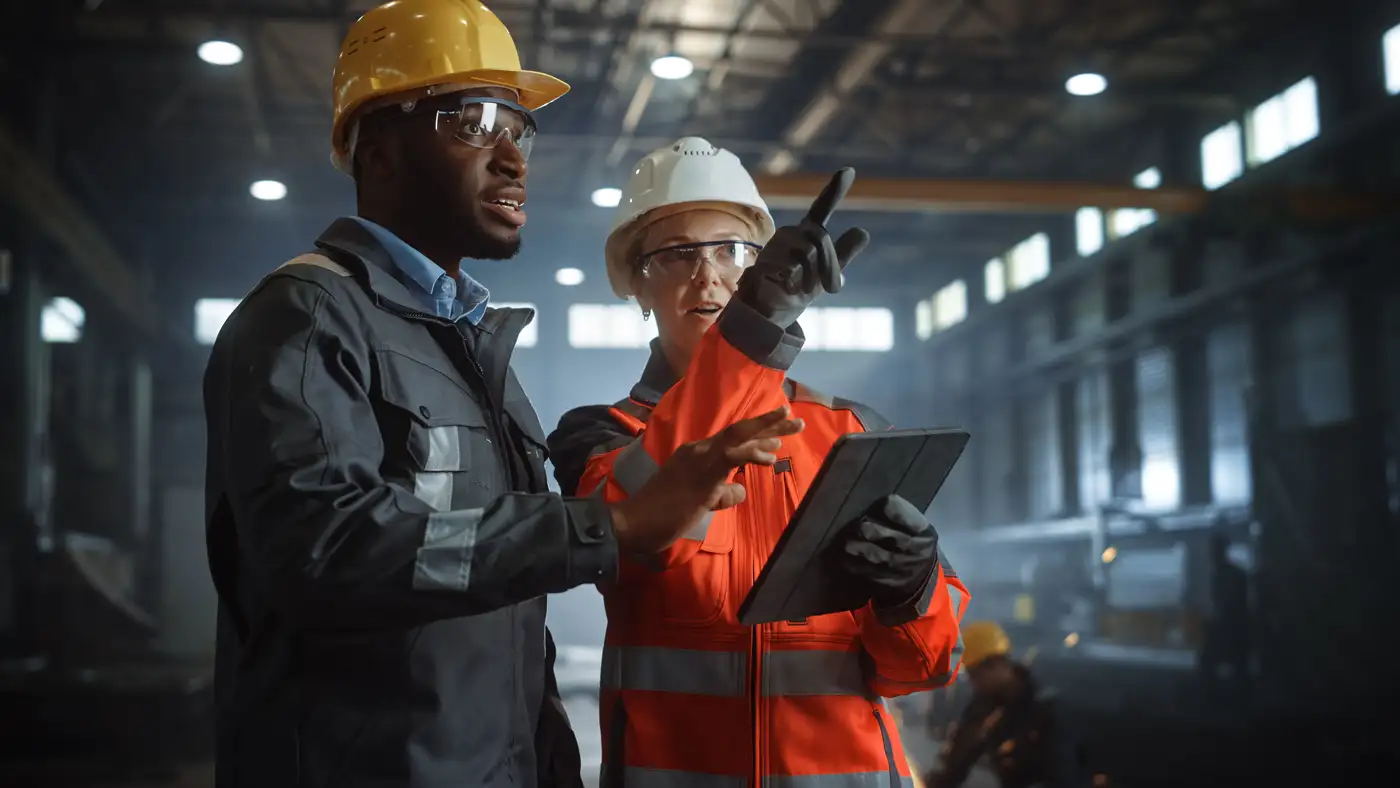 two pleople on the plant floor wearing safety gear - pointing and discussing