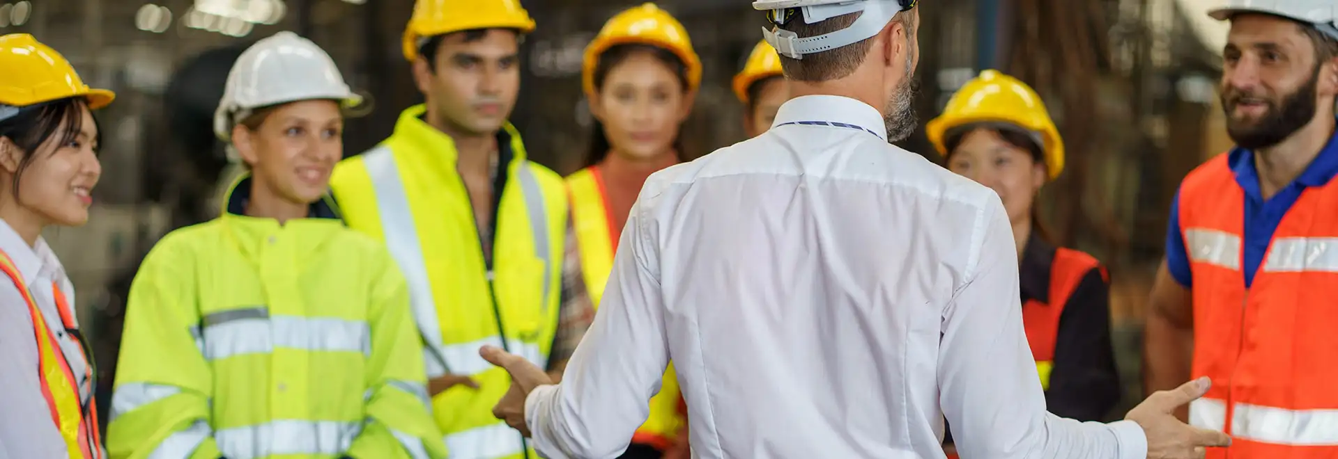person facing away with a white collared shirt speaking to a group all are wearing hard hats