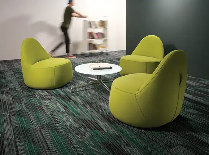 blurred person pushing a cart with a modern seating area in the foreground with green chairs on carpet tile flooring 
