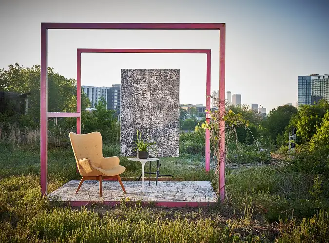 red metal sqaure frame with yellow modern chair with hanging carpet in a nature landscape with buildings in the background