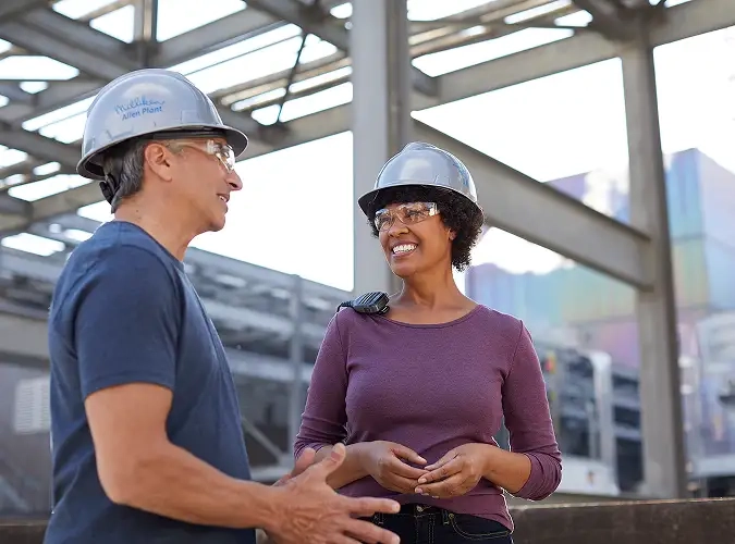 Smiling man and woman wearing hard hats in fornt of industrial site