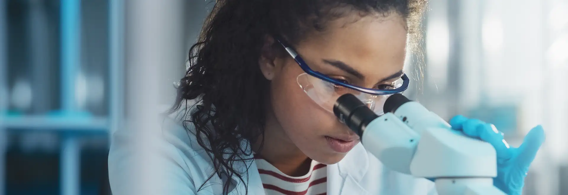 Chemist looing through microscope wearing saftey glasses and gloves