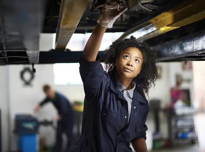 woman in workwear under a lifted car in a repair garage